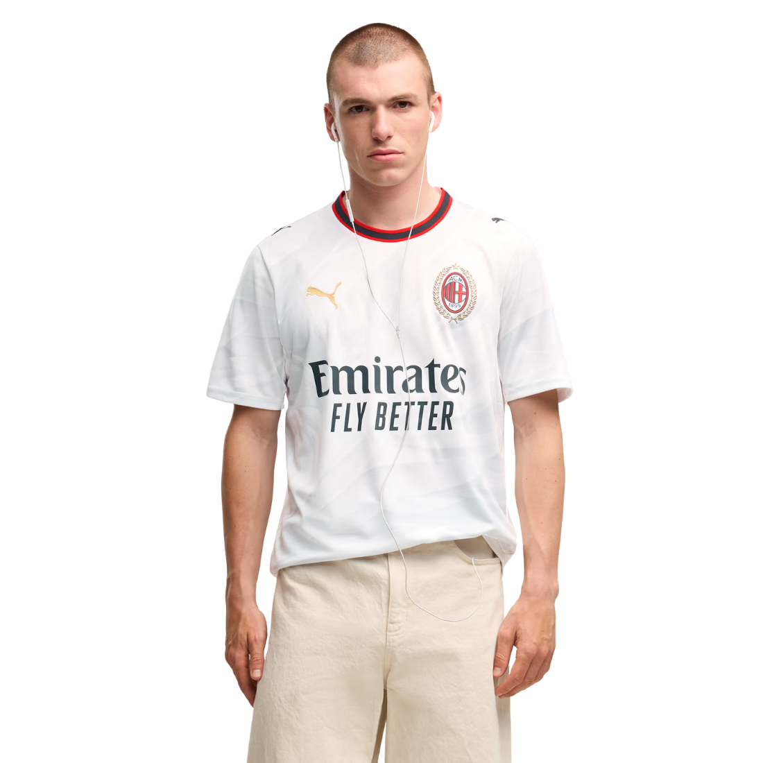 A male model wearing a white Puma AC Milan soccer jersey with the Emirates Fly Better sponsor logo, paired with beige trousers, against a plain white background.