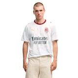 A male model wearing a white Puma AC Milan soccer jersey with the Emirates Fly Better sponsor logo, paired with beige trousers, against a plain white background.