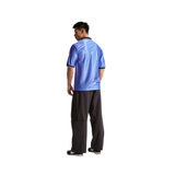 A man shown from the back wearing a shiny light blue referee shirt with black trim and dark gray pants, standing against a plain white background.