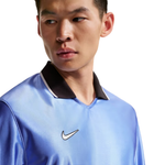 A close-up of a man wearing a light blue Nike athletic jersey with a black collar and a white embroidered swoosh logo on the chest, set against a plain white background.