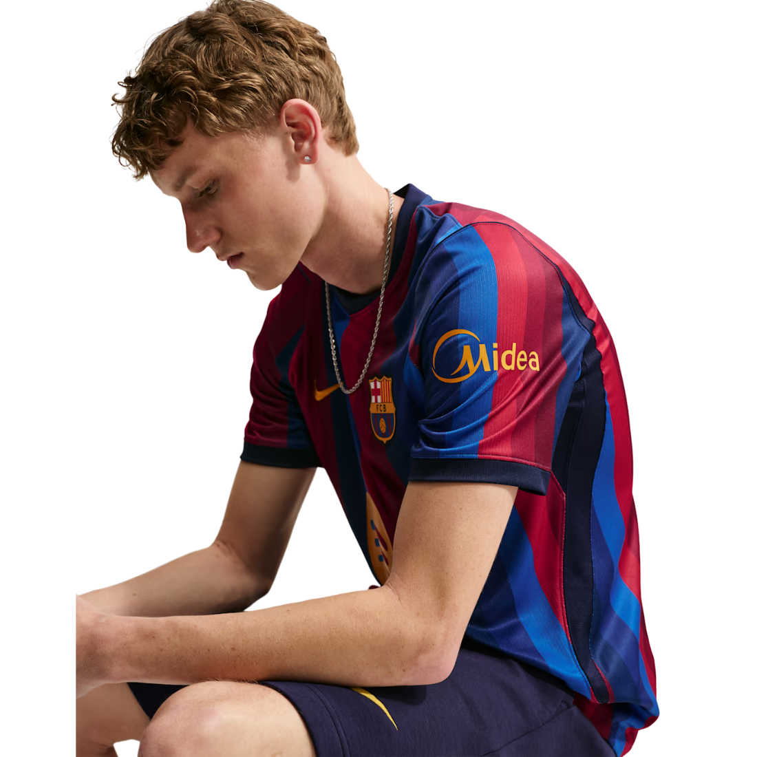 A young man wearing a red and blue striped FC Barcelona soccer jersey with a Midea sponsor logo and Nike swoosh, paired with navy shorts, set against a plain white background.