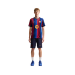 A male model stands against a plain white background wearing a FC Barcelona home soccer jersey, navy shorts, white socks, and blue cleats. The jersey features vertical red and blue stripes with a yellow circular logo on the chest, and a visible Nike swoosh.