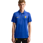 A young man modeling a blue Chelsea FC Nike polo shirt with yellow accents, paired with black shorts, against a plain white background.