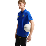 A male model wearing a blue Nike polo shirt with a yellow swoosh and Chelsea crest, holding a white soccer ball over his shoulder against a white background.