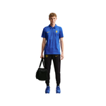 A male model stands against a plain white background wearing a blue Nike polo shirt with a yellow crest, black track pants with a yellow Nike logo, blue sneakers, and holding a black duffel bag.