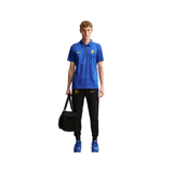 A male model stands against a plain white background wearing a blue Nike polo shirt with a yellow crest, black track pants with a yellow Nike logo, blue sneakers, and holding a black duffel bag.