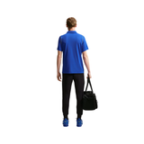 A man shown from behind wearing a bright blue short-sleeve polo shirt, black athletic pants, and blue sneakers, while carrying a black duffel bag against a plain white background.