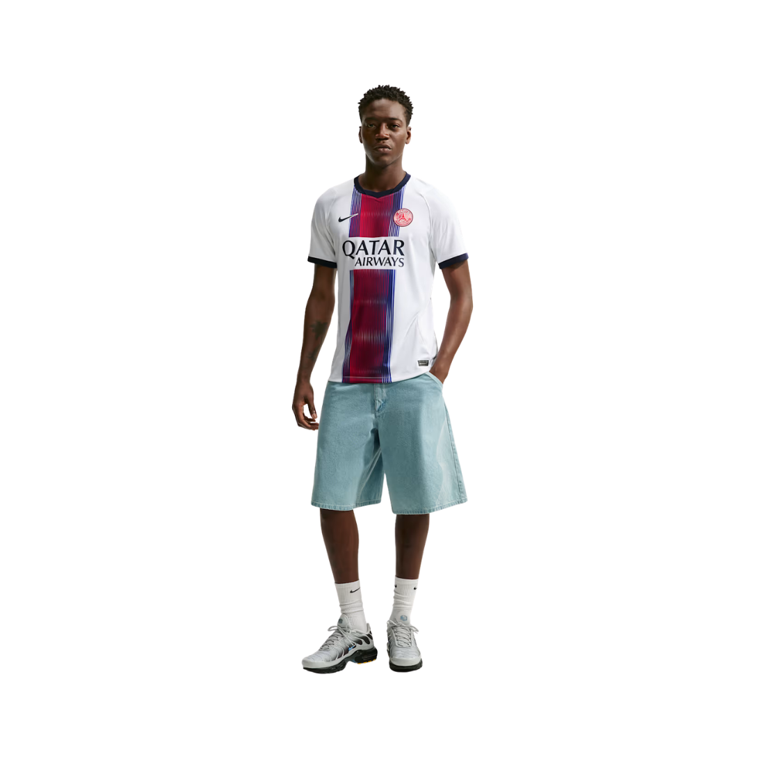 A male model stands against a plain white background wearing a white FC Barcelona soccer jersey with a vertical red and blue stripe, light blue shorts, white socks, and white and black sneakers.