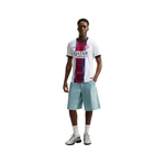 A male model stands against a plain white background wearing a white FC Barcelona soccer jersey with a vertical red and blue stripe, light blue shorts, white socks, and white and black sneakers.