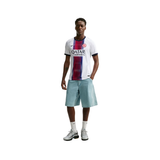 A male model stands against a plain white background wearing a white FC Barcelona soccer jersey with a vertical red and blue stripe, light blue shorts, white socks, and white and black sneakers.