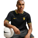 A man wearing a black Nike Chelsea soccer jersey with gold trim and crest, holding a white soccer ball against a white studio background.