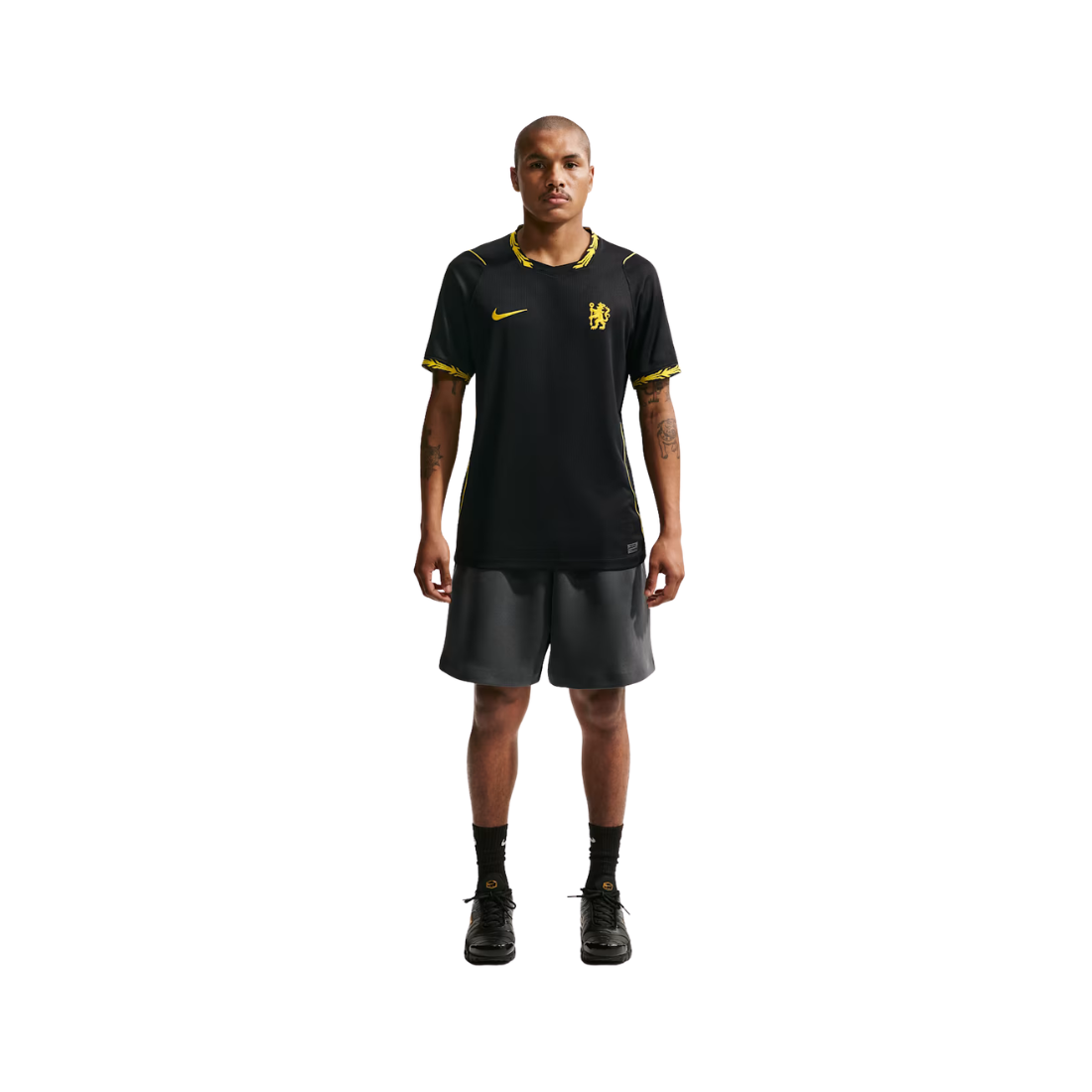 A male model stands against a solid white background wearing a black Nike soccer jersey with yellow accents and dark gray shorts.