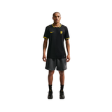 A male model stands against a solid white background wearing a black Nike soccer jersey with yellow accents and dark gray shorts.
