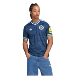 A man wearing a blue Adidas football jersey with white stripes and a crest, paired with blue jeans, against a plain white background.