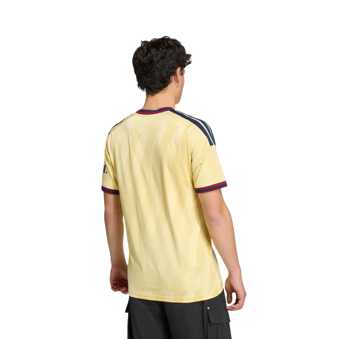 A man viewed from the back wearing a pale yellow short-sleeve soccer jersey with navy and burgundy stripes on the shoulders and collar, paired with black cargo pants, against a plain white background.