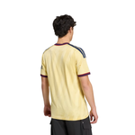 A man viewed from the back wearing a pale yellow short-sleeve soccer jersey with navy and burgundy stripes on the shoulders and collar, paired with black cargo pants, against a plain white background.