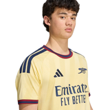 A young man wearing a pale yellow Arsenal soccer jersey with navy and red trim, featuring the Adidas logo and Emirates Fly Better sponsor text, set against a plain white background.