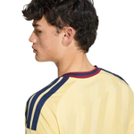 A close-up side profile of a young man wearing a pale yellow soccer jersey with navy blue shoulder stripes and a red and navy collar, against a plain white background.