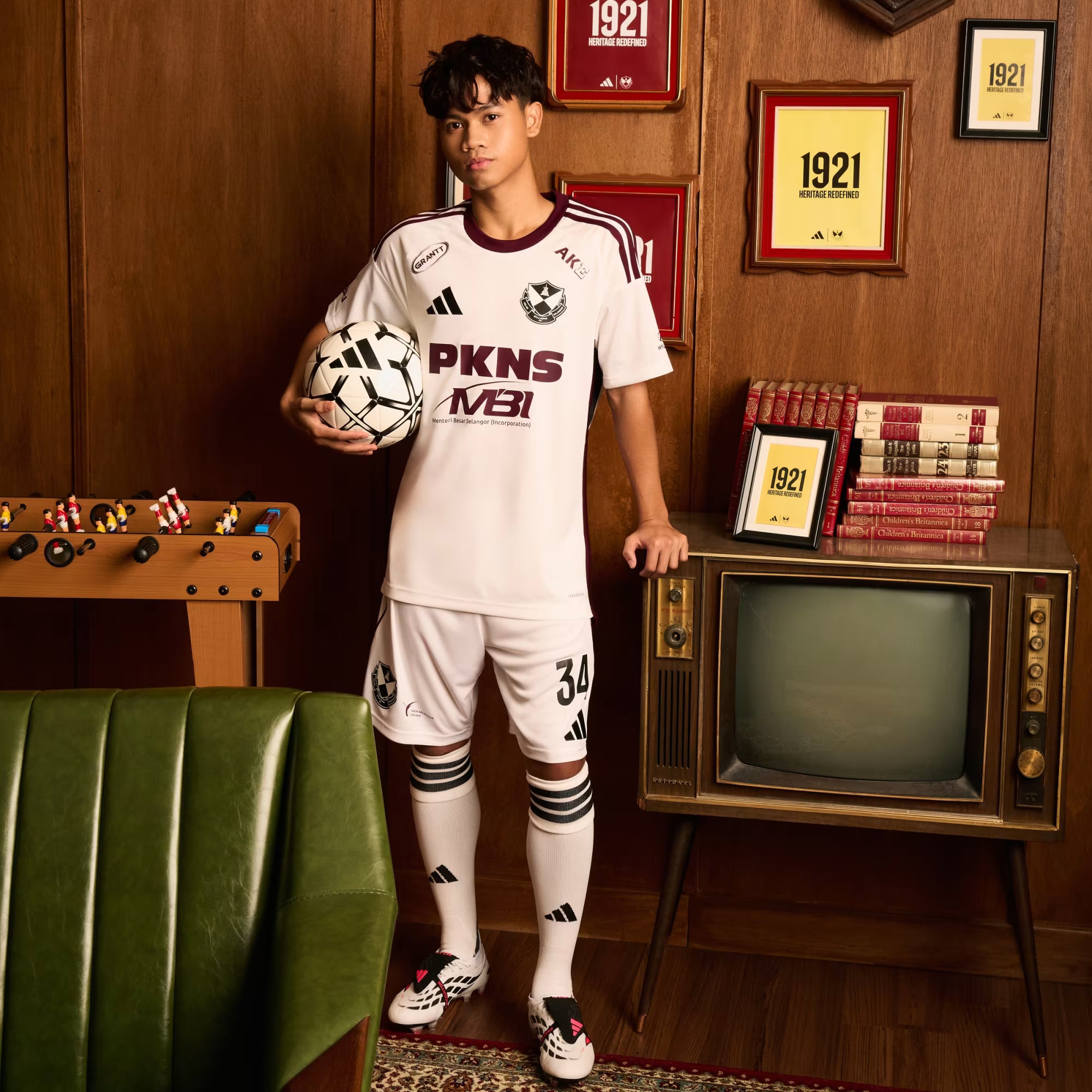 A man modeling a white Adidas soccer kit with maroon accents, holding a soccer ball in a retro-themed room with wood-paneled walls, framed certificates, a green bench, and a vintage television.
