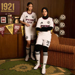 Two female models wearing white PKNNS IVNS soccer jerseys, shorts, and knee-high socks with black stripes, posed in a themed indoor setting with vintage 1921 decor, soccer balls, and a striped sofa.