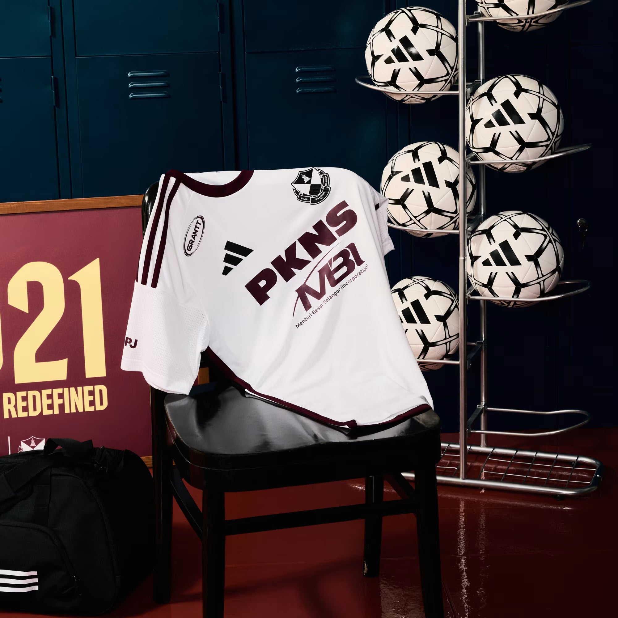 A white soccer jersey with maroon trim and 'PKNS' and 'AVEN' branding, draped over a black chair in a locker room setting with soccer balls on a rack and a black bag nearby.